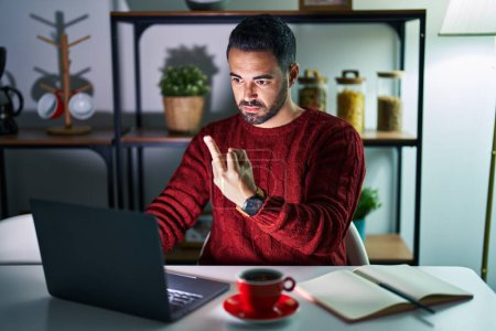 Foto de Young hispanic man with beard using computer laptop at night at home showing middle finger, impolite and rude fuck off expression - Imagen libre de derechos