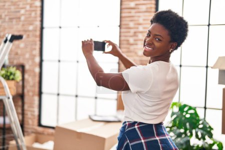 Téléchargez les photos : Femme afro-américaine souriant confiant faire la photo par le smartphone à la nouvelle maison - en image libre de droit