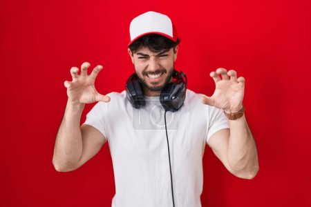 Téléchargez les photos : Homme hispanique avec barbe portant chapeau de joueur et écouteurs souriant drôle faisant griffe geste comme chat, expression agressive et sexy - en image libre de droit
