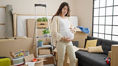 Foto de Mujer embarazada joven mirando a su alrededor con expresión seria tocando el vientre en el nuevo hogar - Imagen libre de derechos
