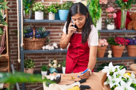 Téléchargez les photos : Jeune belle hispanique femme fleuriste parler sur smartphone écriture sur enveloppe lettre à la boutique de fleurs - en image libre de droit