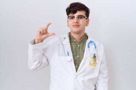 Photo for Young non binary man wearing doctor uniform and stethoscope smiling and confident gesturing with hand doing small size sign with fingers looking and the camera. measure concept. - Royalty Free Image