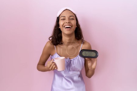 Foto de Mujer hispana joven vistiendo camisón con despertador sonriendo y riendo en voz alta porque divertida broma loca. - Imagen libre de derechos