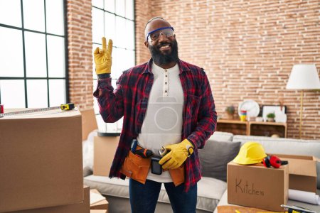 Foto de Hombre afroamericano trabajando en la renovación del hogar sonriendo con la cara feliz guiñando el ojo a la cámara haciendo señal de victoria. número dos. - Imagen libre de derechos