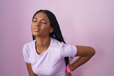 Téléchargez les photos : Femme afro-américaine avec des tresses debout sur fond rose souffrant de maux de dos, toucher le dos avec la main, douleur musculaire - en image libre de droit