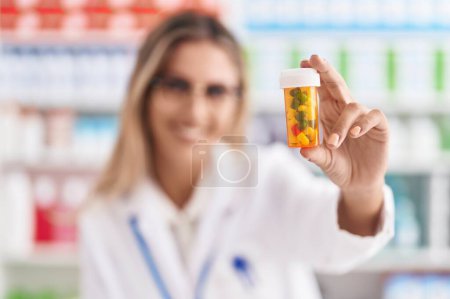 Foto de Joven mujer rubia farmacéutico sonriendo confiado sosteniendo pastillas botella en la farmacia - Imagen libre de derechos