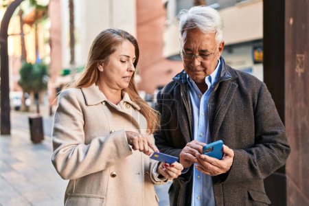 Téléchargez les photos : Couple homme / femme d'âge moyen utilisant smartphone et carte de crédit dans la rue - en image libre de droit