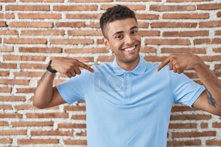 Photo for Brazilian young man standing over brick wall looking confident with smile on face, pointing oneself with fingers proud and happy. - Royalty Free Image