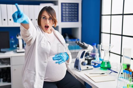 Photo for Pregnant woman working at scientist laboratory pointing with finger surprised ahead, open mouth amazed expression, something on the front - Royalty Free Image