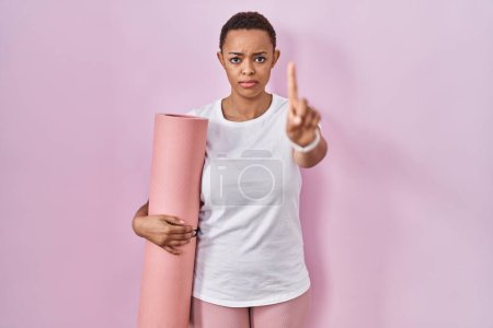 Foto de Hermosa mujer afroamericana sosteniendo esterilla de yoga señalando con el dedo hacia arriba y la expresión enojada, sin mostrar ningún gesto - Imagen libre de derechos