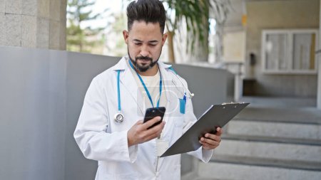 Photo for Young latin man doctor using smartphone holding clipboard at hospital - Royalty Free Image