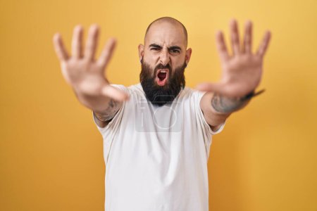Foto de Joven hombre hispano con barba y tatuajes de pie sobre fondo amarillo haciendo gesto de stop con palmas de las manos, expresión de enojo y frustración - Imagen libre de derechos