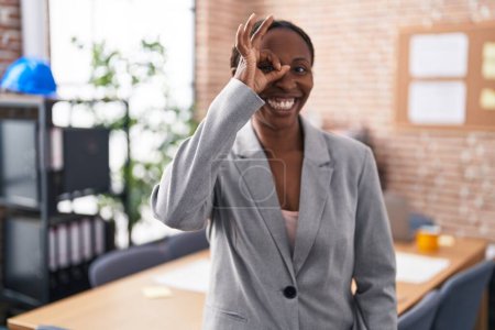 Téléchargez les photos : Afro-américaine femme au bureau souriant heureux faire ok signe avec main sur oeil regarder à travers les doigts - en image libre de droit