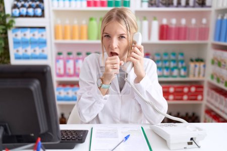 Foto de Mujer caucásica joven que trabaja en la farmacia hablando por teléfono sintiéndose mal y tosiendo como síntoma de resfriado o bronquitis. concepto de atención sanitaria. - Imagen libre de derechos