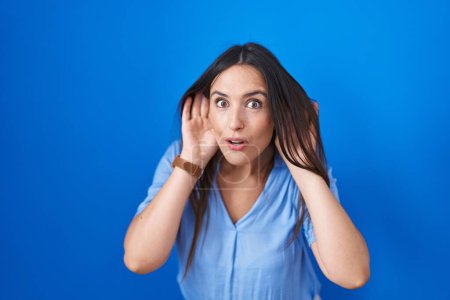 Photo for Young brunette woman standing over blue background trying to hear both hands on ear gesture, curious for gossip. hearing problem, deaf - Royalty Free Image