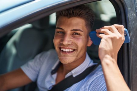 Photo for Young hispanic man listening audio message by smartphone sitting on car at street - Royalty Free Image