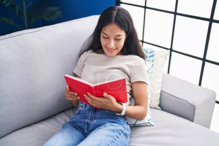 Photo for Young beautiful hispanic woman reading book sitting on sofa at home - Royalty Free Image