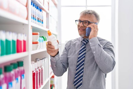 Téléchargez les photos : Middle age grey-haired man customer talking on smartphone holding sunscreen bottle at laboratory - en image libre de droit