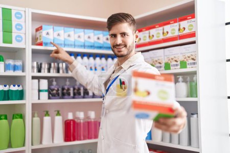 Téléchargez les photos : Jeune homme caucasien pharmacien souriant confiant tenant paquet de vitamines à la pharmacie - en image libre de droit