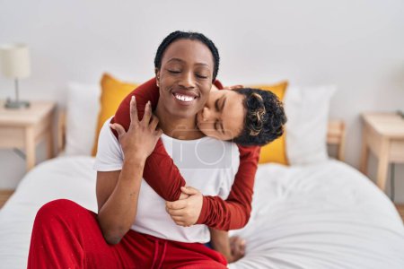 Foto de Afro-americano madre e hijo abrazándose y besándose sentado en la cama en el dormitorio - Imagen libre de derechos