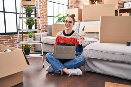 Photo for Young hispanic girl sitting on the floor at new home with laptop smiling happy pointing with hand and finger to the side - Royalty Free Image