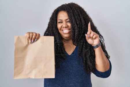 Foto de Más tamaño mujer hispana sosteniendo llevar bolsa de papel sorprendido con una idea o pregunta señalando el dedo con la cara feliz, número uno - Imagen libre de derechos