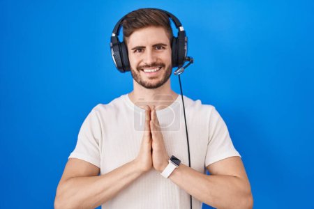 Foto de Hombre hispano con barba escuchando música usando auriculares orando con las manos juntas pidiendo perdón sonriendo confiado. - Imagen libre de derechos