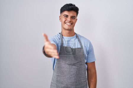 Foto de Joven hispano vistiendo delantal sobre fondo blanco sonriente amistoso ofreciendo apretón de manos como saludo y bienvenida. negocios exitosos. - Imagen libre de derechos