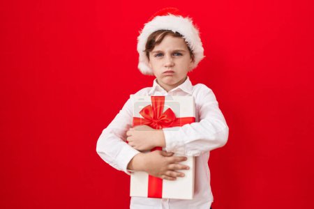 Téléchargez les photos : Petit garçon caucasien portant un chapeau de Noël et tenant des cadeaux sceptiques et nerveux, fronçant les sourcils contrariés à cause de problèmes. personne négative. - en image libre de droit