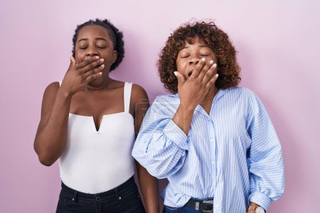 Téléchargez les photos : Deux femmes africaines debout sur fond rose s'ennuient à bâiller fatigué couvrant la bouche avec la main. agitation et somnolence. - en image libre de droit