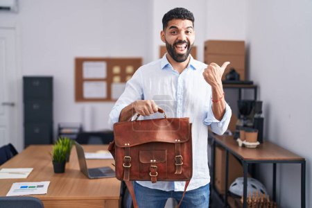 Téléchargez les photos : Homme hispanique avec barbe travaillant au bureau tenant la mallette pointant le pouce vers le côté souriant heureux avec la bouche ouverte - en image libre de droit