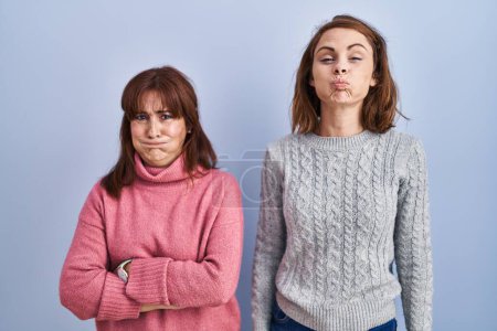 Photo for Mother and daughter standing over blue background puffing cheeks with funny face. mouth inflated with air, crazy expression. - Royalty Free Image