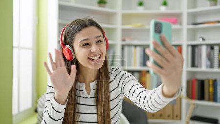 Téléchargez les photos : Jeune belle étudiante hispanique souriante confiante ayant un appel vidéo à la bibliothèque de l'université - en image libre de droit