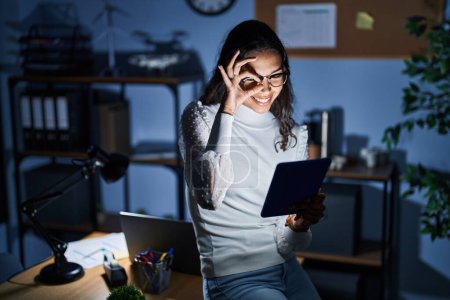 Photo for Young brazilian woman using touchpad at night working at the office doing ok gesture with hand smiling, eye looking through fingers with happy face. - Royalty Free Image