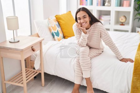 Foto de Joven hermosa mujer hispana sonriendo confiada sentada en la cama en el dormitorio - Imagen libre de derechos