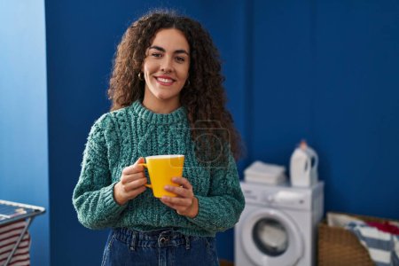 Téléchargez les photos : Young beautiful hispanic woman drinking coffee waiting for washing machine at laundry room - en image libre de droit