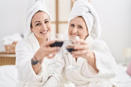 Téléchargez les photos : Mother and daughter wearing bathrobe drinking glass of wine at bedroom - en image libre de droit