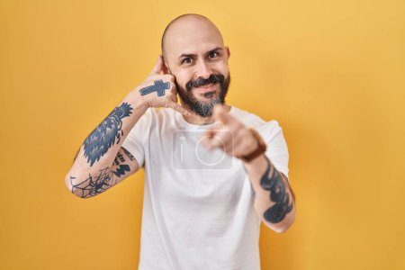 Téléchargez les photos : Jeune homme hispanique avec des tatouages debout sur fond jaune souriant faire parler au téléphone geste et vous pointant du doigt. Appelle-moi. - en image libre de droit