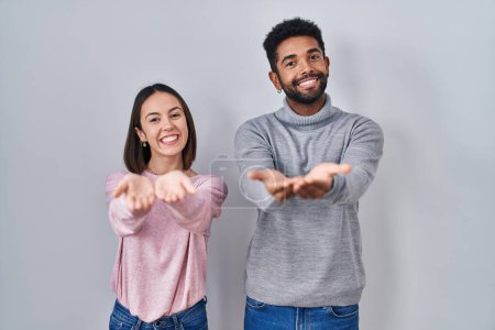 Téléchargez les photos : Jeune couple hispanique debout ensemble souriant avec les mains paumes ensemble recevoir ou donner geste. maintien et protection - en image libre de droit