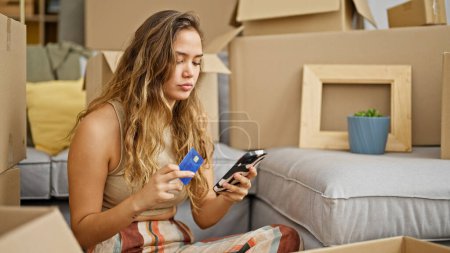Foto de Joven hermosa mujer hispana de compras con teléfono inteligente y tarjeta de crédito sentado en el piso en el nuevo hogar - Imagen libre de derechos