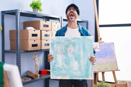 Foto de Hombre hispano con barba sosteniendo lienzo en el estudio enojado y loco gritando frustrado y furioso, gritando con ira mirando hacia arriba. - Imagen libre de derechos