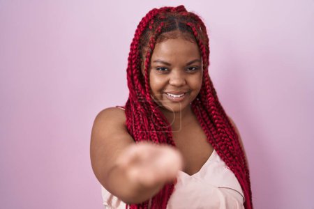 Foto de Mujer afroamericana con cabello trenzado de pie sobre fondo rosa sonriente alegre que ofrece la palma de la mano dando asistencia y aceptación. - Imagen libre de derechos