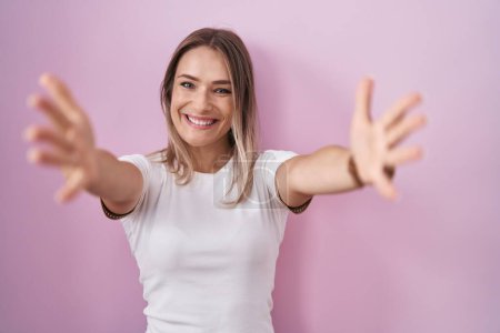 Photo for Blonde caucasian woman standing over pink background looking at the camera smiling with open arms for hug. cheerful expression embracing happiness. - Royalty Free Image