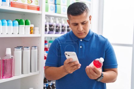 Foto de Joven cliente latino usando el teléfono inteligente que sostiene el frasco de medicina en la farmacia - Imagen libre de derechos