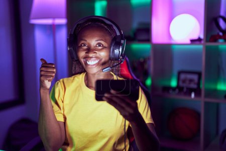 Téléchargez les photos : Femme afro-américaine jouant à des jeux vidéo avec smartphone souriant heureux et positif, pouce levé faire excellent et signe d'approbation - en image libre de droit