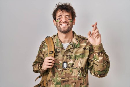 Foto de Joven hispano vistiendo uniforme camuflaje ejército gesto cruzado sonriendo con esperanza y los ojos cerrados. suerte y concepto supersticioso. - Imagen libre de derechos