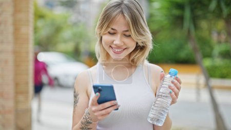 Photo for Young blonde woman using smartphone holding bottle of water at street - Royalty Free Image