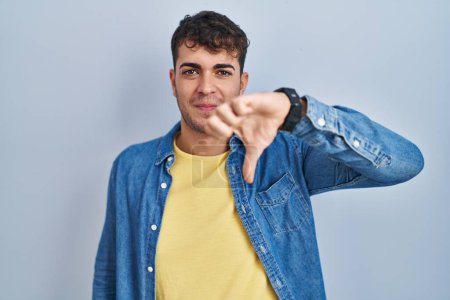 Téléchargez les photos : Young hispanic man standing over blue background looking unhappy and angry showing rejection and negative with thumbs down gesture. bad expression. - en image libre de droit