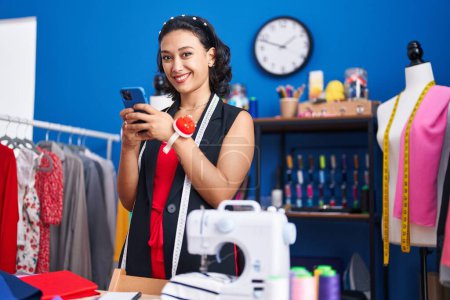 Téléchargez les photos : Jeune belle femme hispanique tailleur souriant confiant en utilisant smartphone à l'usine de vêtements - en image libre de droit