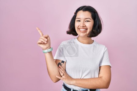 Foto de Mujer hispana joven vistiendo una camiseta blanca casual sobre fondo rosa con una gran sonrisa en la cara, señalando con el dedo de la mano hacia el lado mirando a la cámara. - Imagen libre de derechos